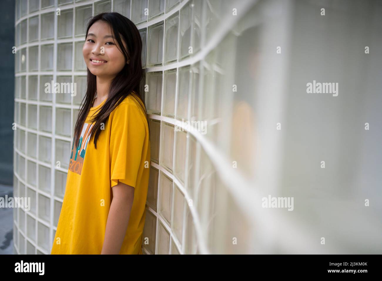 Ritratto di una ragazzina che indossa una maglietta gialla e si trova contro una parete di blocchi di vetro; Hong Kong, Cina Foto Stock