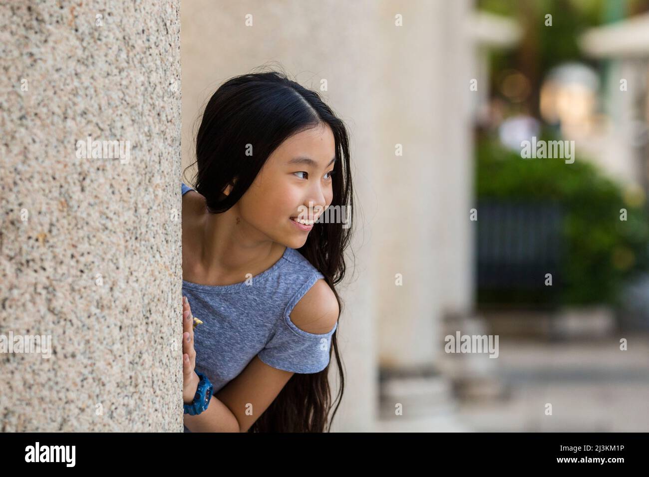 Ritratto all'aperto di una ragazza con capelli lunghi e scuri come lei guarda fuori da dietro il muro e guarda a destra; Stanley, Hong Kong, Cina Foto Stock
