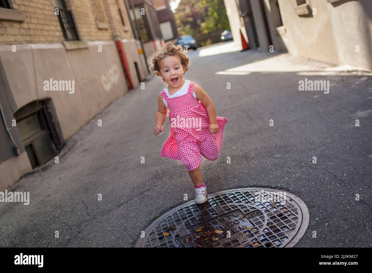 Ragazza preschooler che corre in un vicolo nel suo abito bianco e rosso di Gingham; Toronto, Ontario, Canada Foto Stock