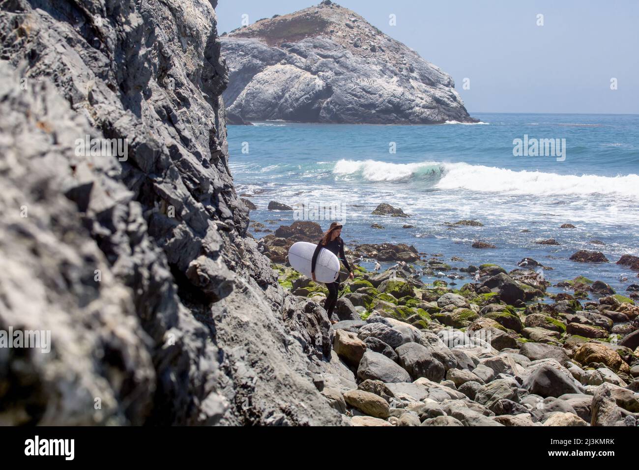 Un surfista attraversa un masso disseminata spiaggia per arrivare alle onde. Foto Stock