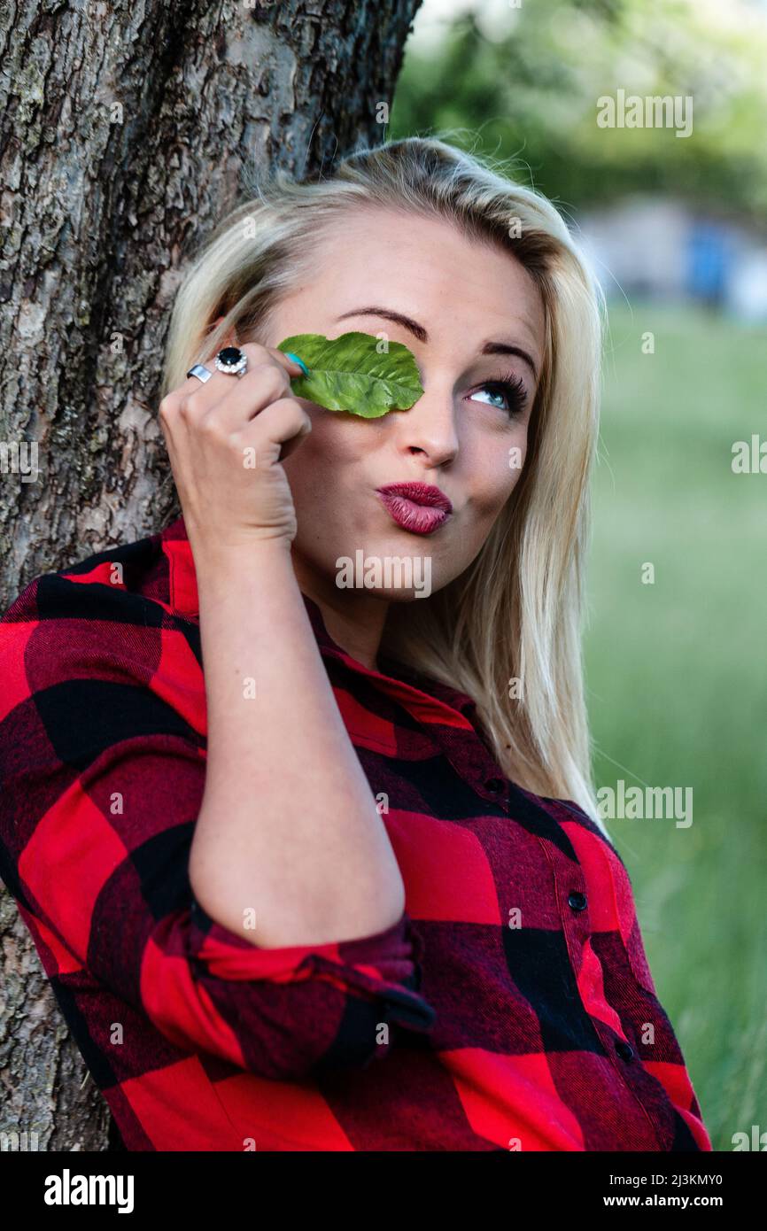 Giovane donna giocosa che copre il suo occhio con una foglia verde guardando in su con un sorriso e le labbra pursed mentre si rilassa contro un tronco di albero in un parco Foto Stock