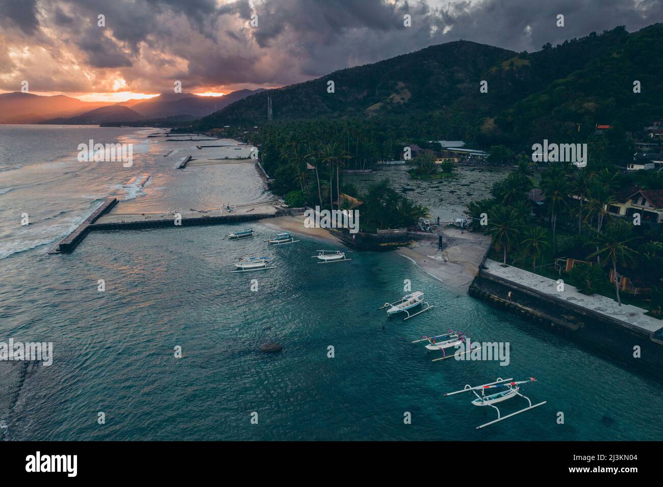 Vista aerea dei canoe jukung ormeggiati a un molo e lungo la riva di Candidasa Beach con luce del sole che scorre attraverso le montagne sotto un grigio c... Foto Stock