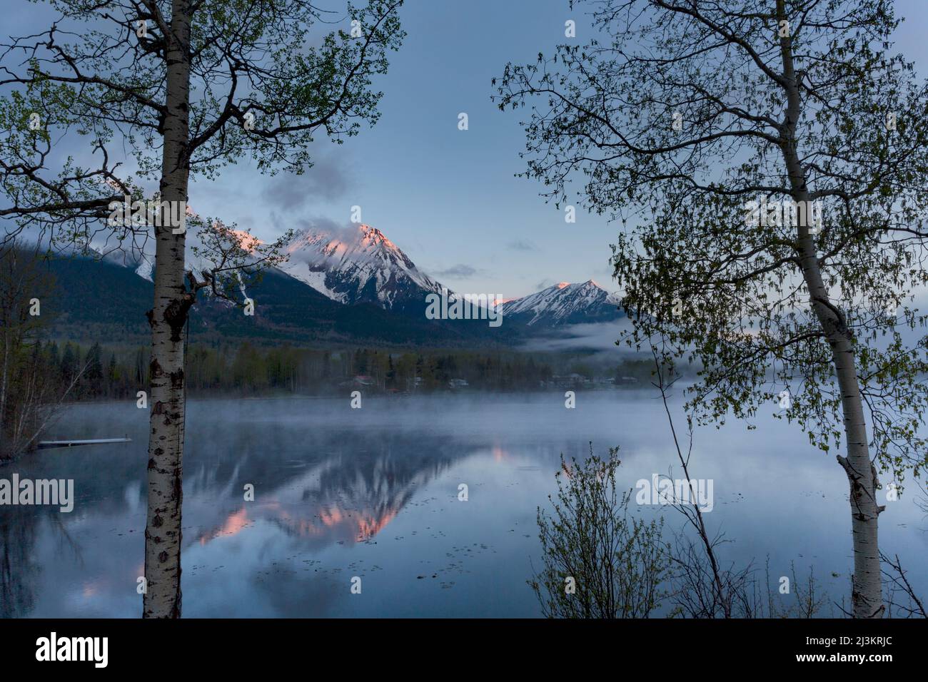 Aspra vetta della Hudson Bay Mountain coperta di neve con nebbia che si innalza dal lago in primo piano all'alba; Smithers, British Columbia, Canada Foto Stock