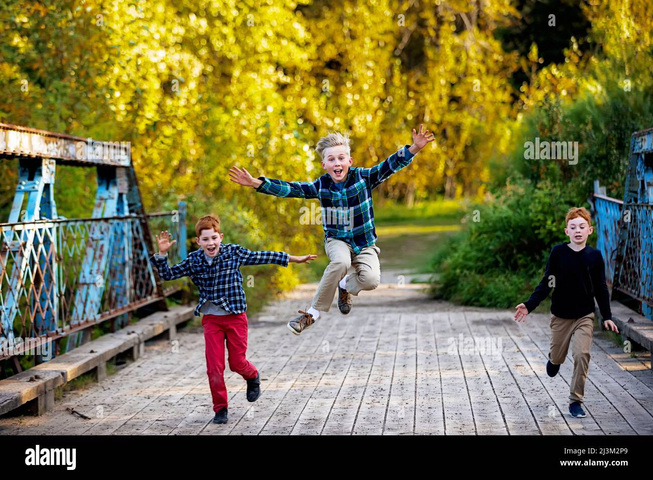 Tre ragazzi che corrono attraverso un ponte in un parco in autunno, con il ragazzo al centro che salta in alto nell'aria; Edmonton, Alberta, Canada Foto Stock