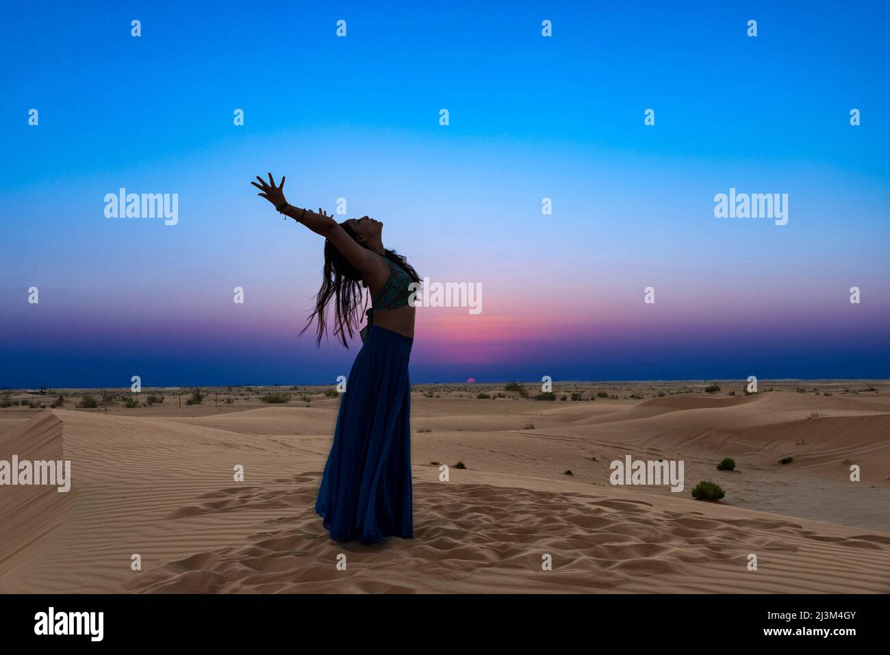 Donna con gonna lunga illuminata dal tramonto sulle sabbie del deserto, in piedi in una posa yoga curva posteriore; Abu Dhabi, Emirati Arabi Uniti Foto Stock