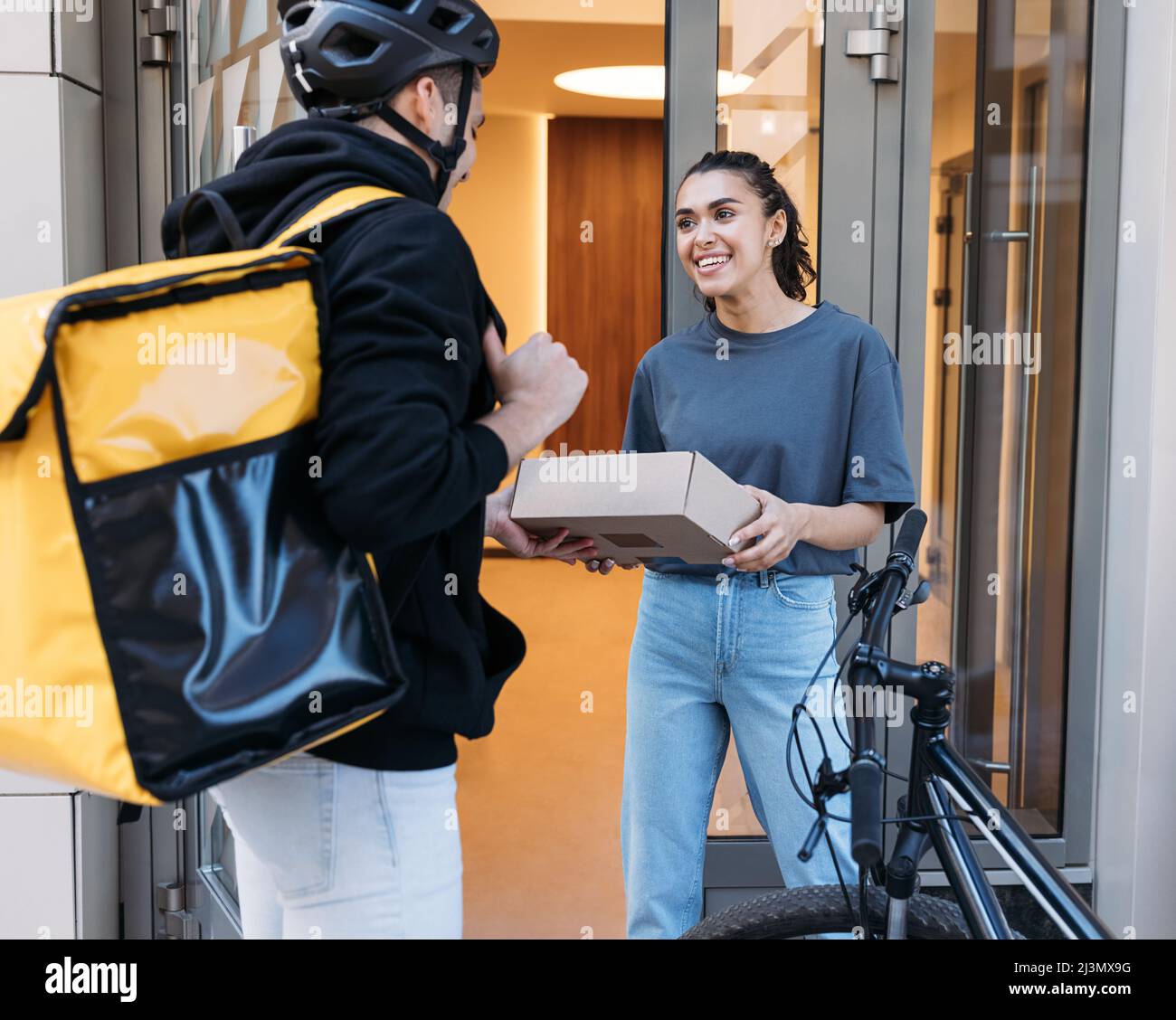 Donna sorridente che riceve un pacco dal corriere. Tipo di consegna con zaino che dà il pacchetto ad un cliente in un edificio di appartamento. Foto Stock