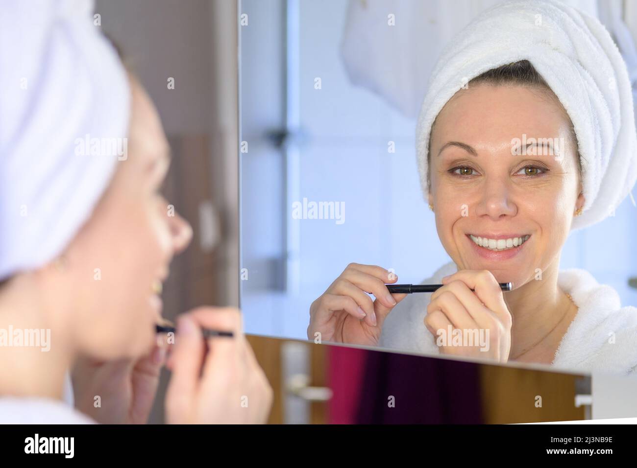 Donna felice con un sorriso vivacious che applica il suo trucco in una vista sopra la spalla al suo riflesso nello specchio della stanza da bagno mentre osserva il camer Foto Stock