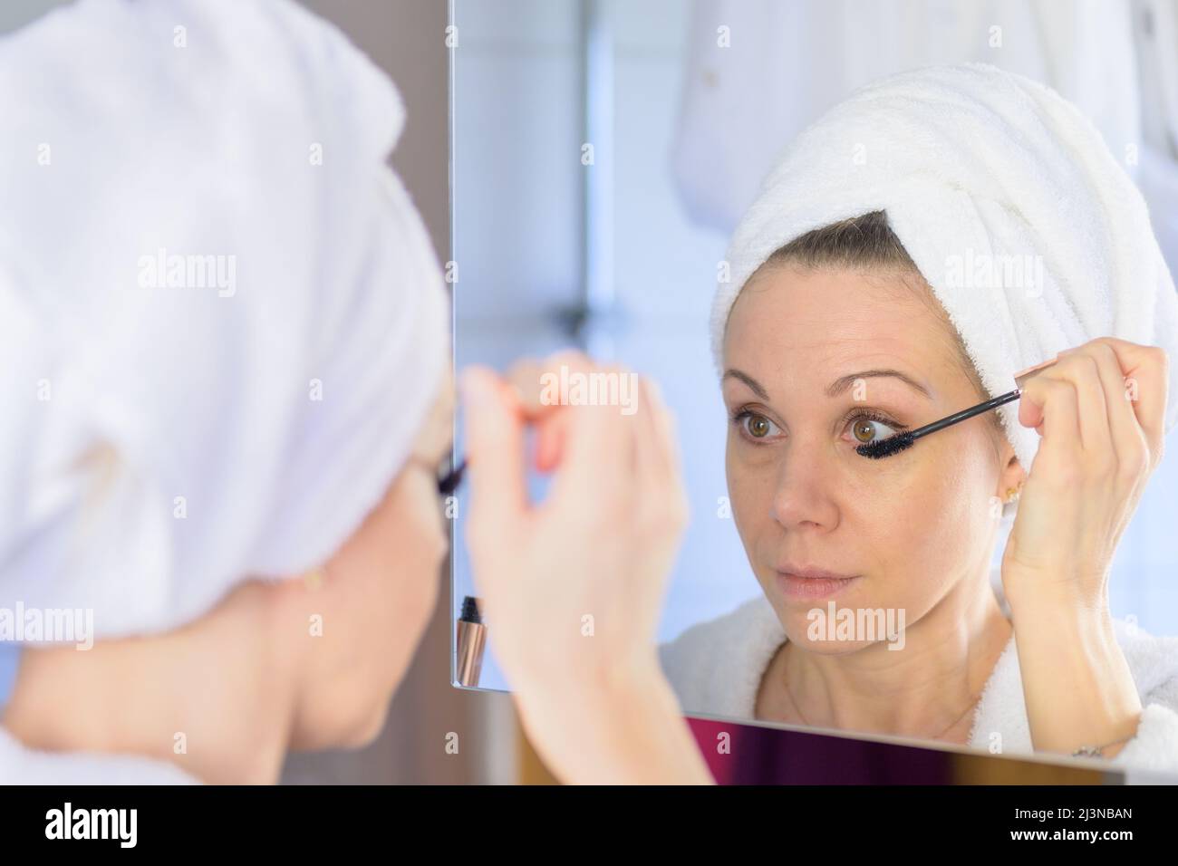 Donna con i suoi capelli legati in un asciugamano che applica mascara nello specchio del bagno in una vista sopra la spalla al suo riflesso Foto Stock