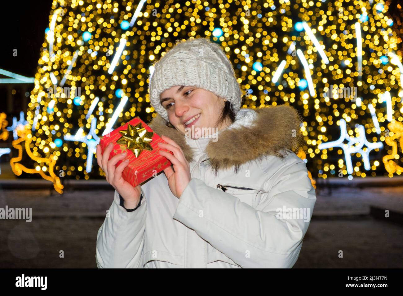 Una donna in abiti bianchi d'inverno si erge fuori da un albero di Natale, contiene una scatola regalo rossa e sorride. Foto Stock