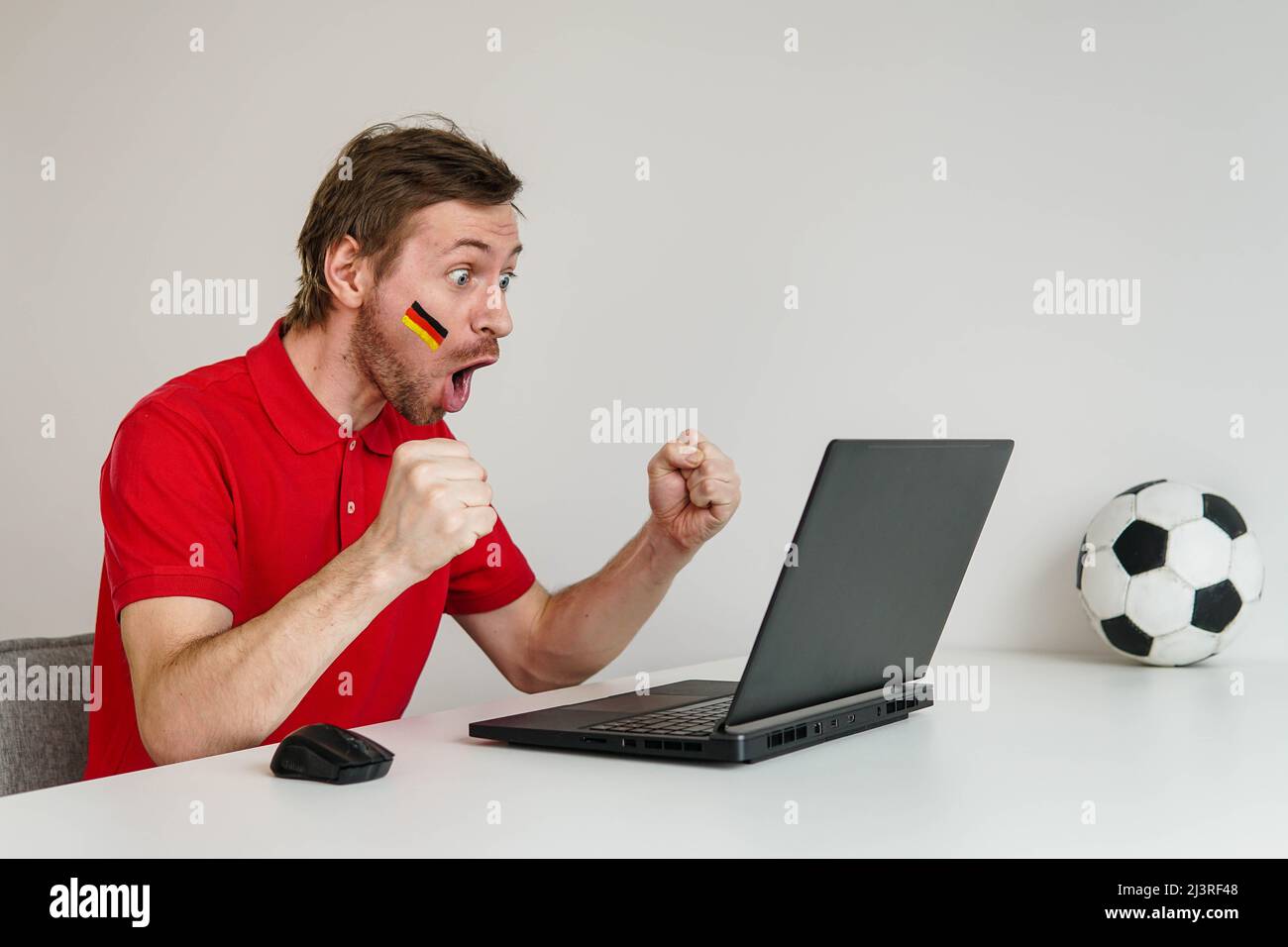 Giovane fan eccitato uomo in camicia rossa sostenere la squadra nazionale tedesca di calcio tenere in mano palla di calcio guardare la tv in diretta streaming sul portatile. Foto Stock