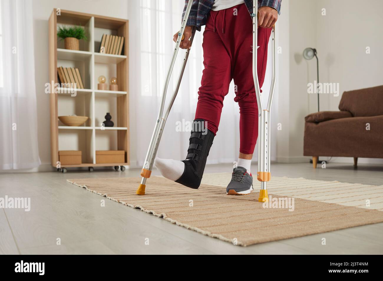 Giovane uomo con piede rotto che indossa un sostegno per le gambe e cammina sulle stampelle a casa Foto Stock