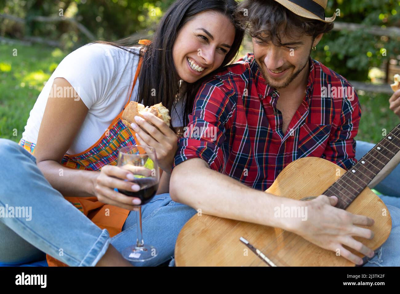 Coppia di giovani che condividono emozioni positive all'aperto divertirsi al picnic. Sta mangiando un panino mentre tiene una chitarra acustica Foto Stock