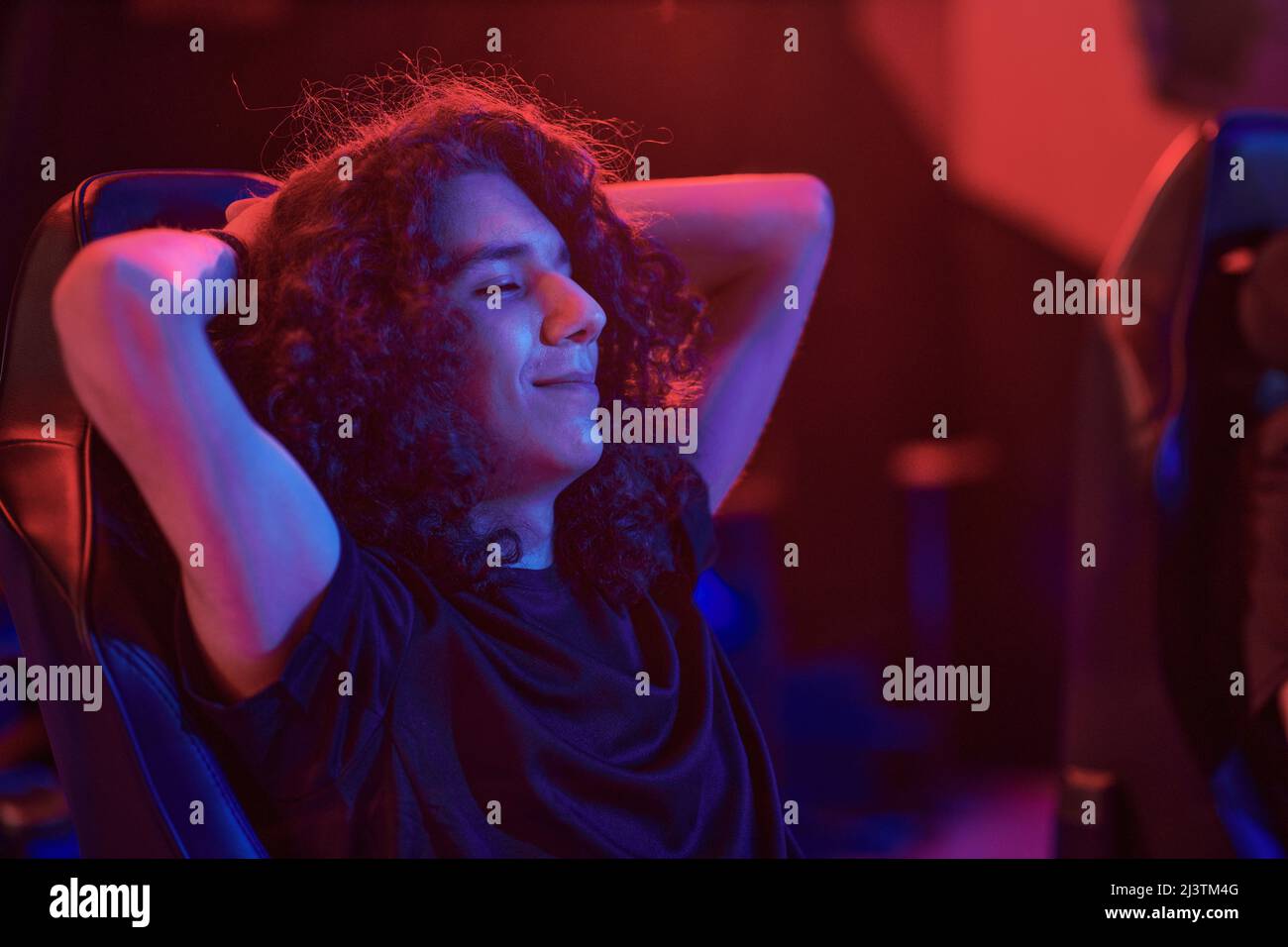Videogamer con capelli ricci lunghi che tengono gli occhi chiusi e tengono le mani dietro la testa mentre si rilassa nella sedia del computer nella barra degli eSport Foto Stock