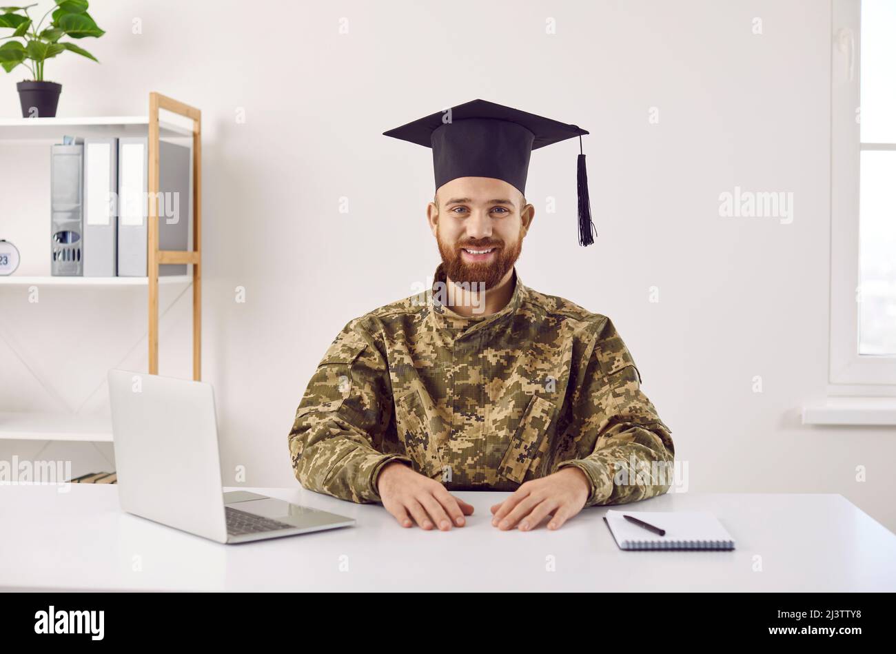 Felice giovane studente in uniforme camouflage e cappello laureato seduto alla scrivania e sorridente Foto Stock