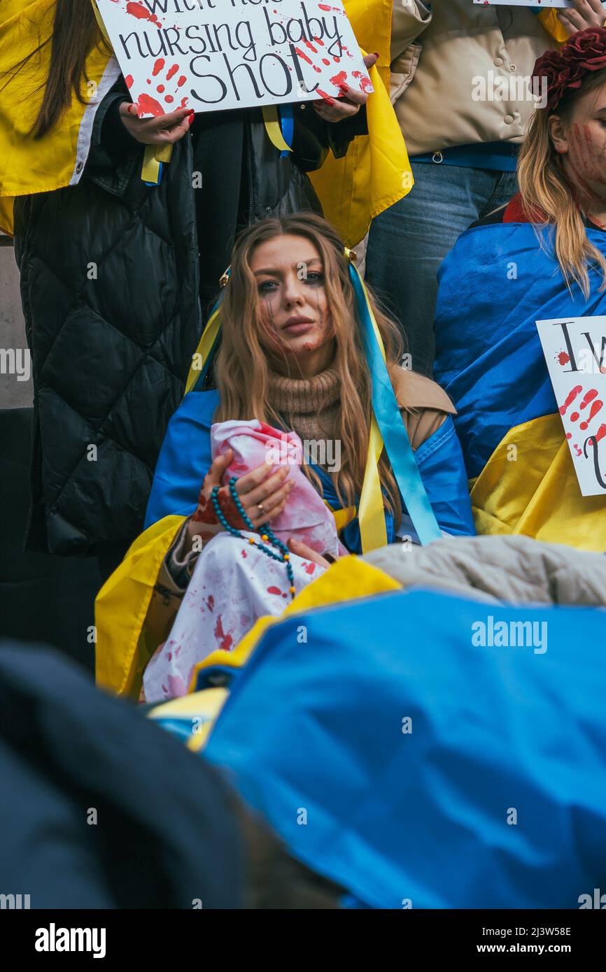 Una donna pro-Ucraina siede per le strade di una dimostrazione con una bandiera Ucraina intorno al suo corpo e tenendo un bambino coperto da un foglio di sangue di guerra Foto Stock