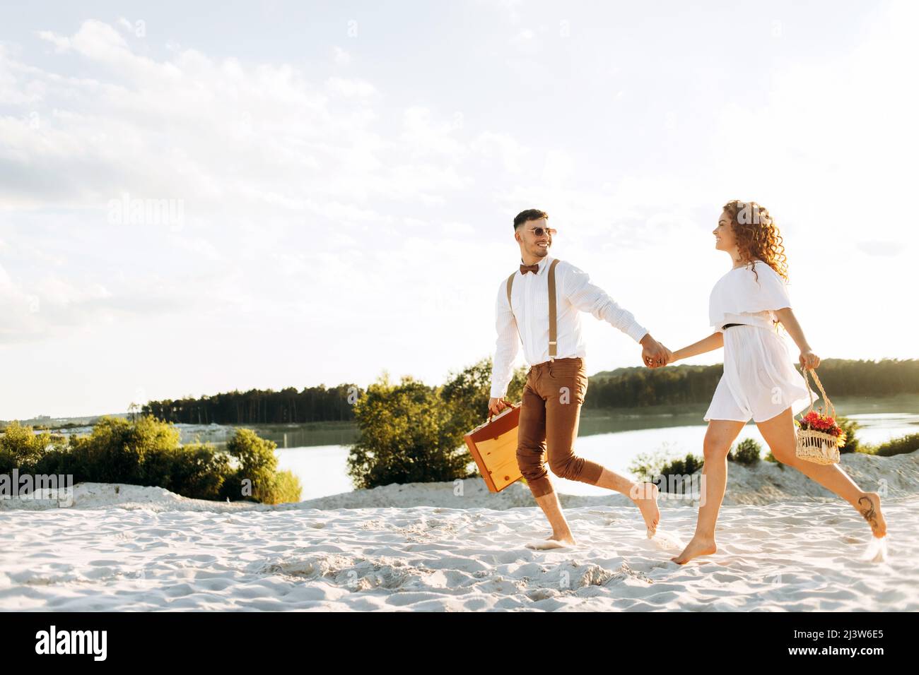 Coppia felice correre sulla sabbia, tramonto, coppia elegante con una valigia e fiori in un cestino. Foto Stock
