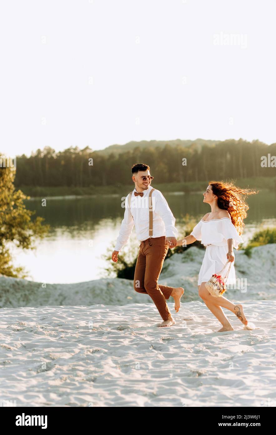 Coppia felice correre sulla sabbia, tramonto, coppia elegante con una valigia e fiori in un cestino. Foto Stock