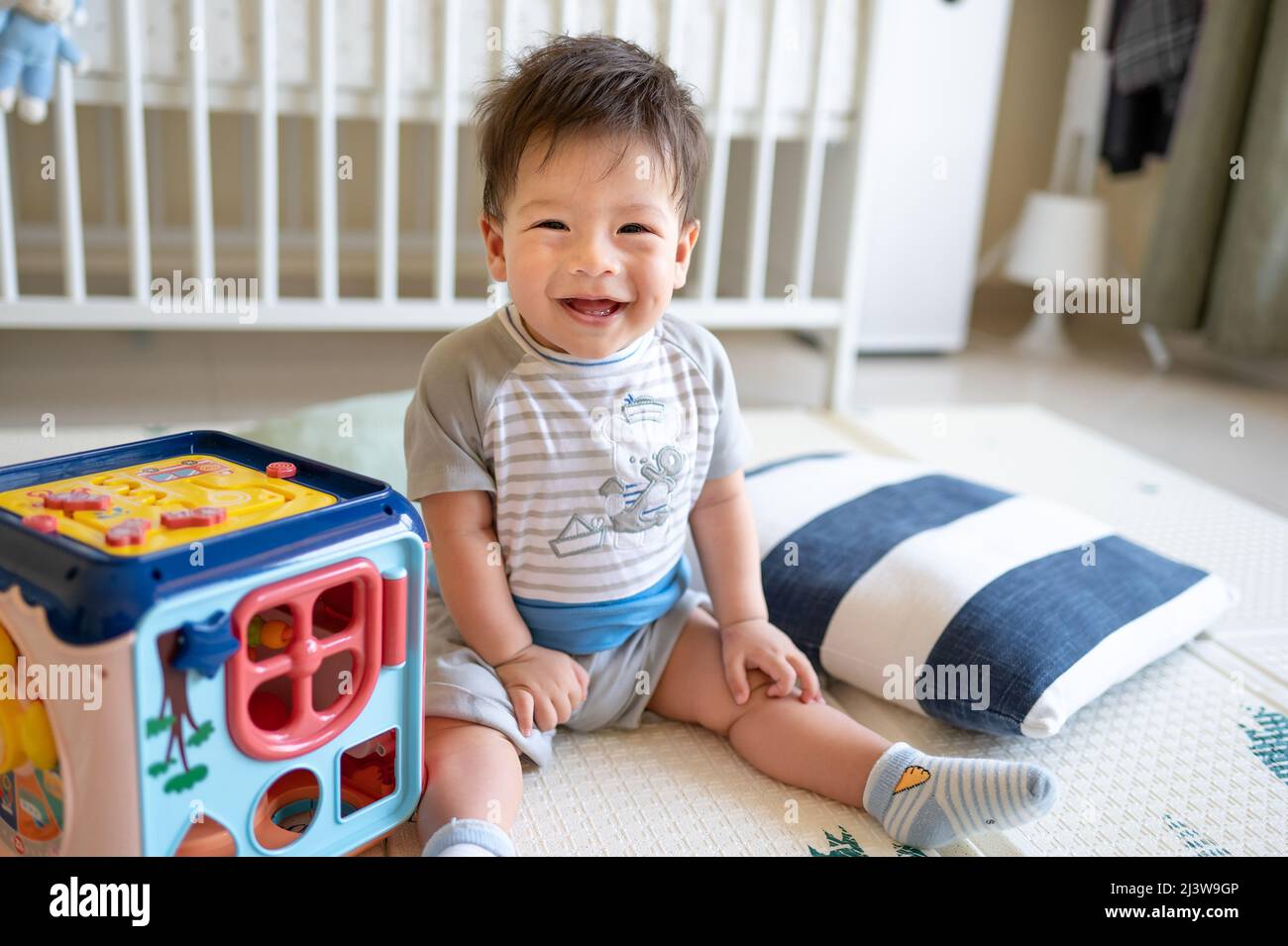 Adorabile bambino di 8 mesi di razza mista sorridendo giocando con box attività in camera da letto mentre si siede sul pavimento coperto da un tappetino gioco di gomma Foto Stock