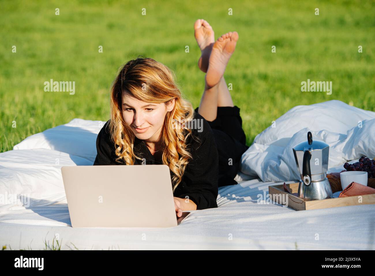 Donna ben riposata che lavora su un computer portatile in un letto sotto un cielo aperto. Su un prato in campagna. Tè e frutta su una tavola accanto a lei. Foto Stock