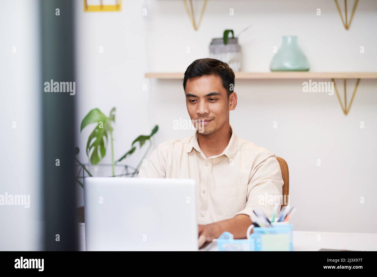 Un giovane uomo asiatico siede alla sua scrivania e digita sul suo computer portatile al lavoro Foto Stock