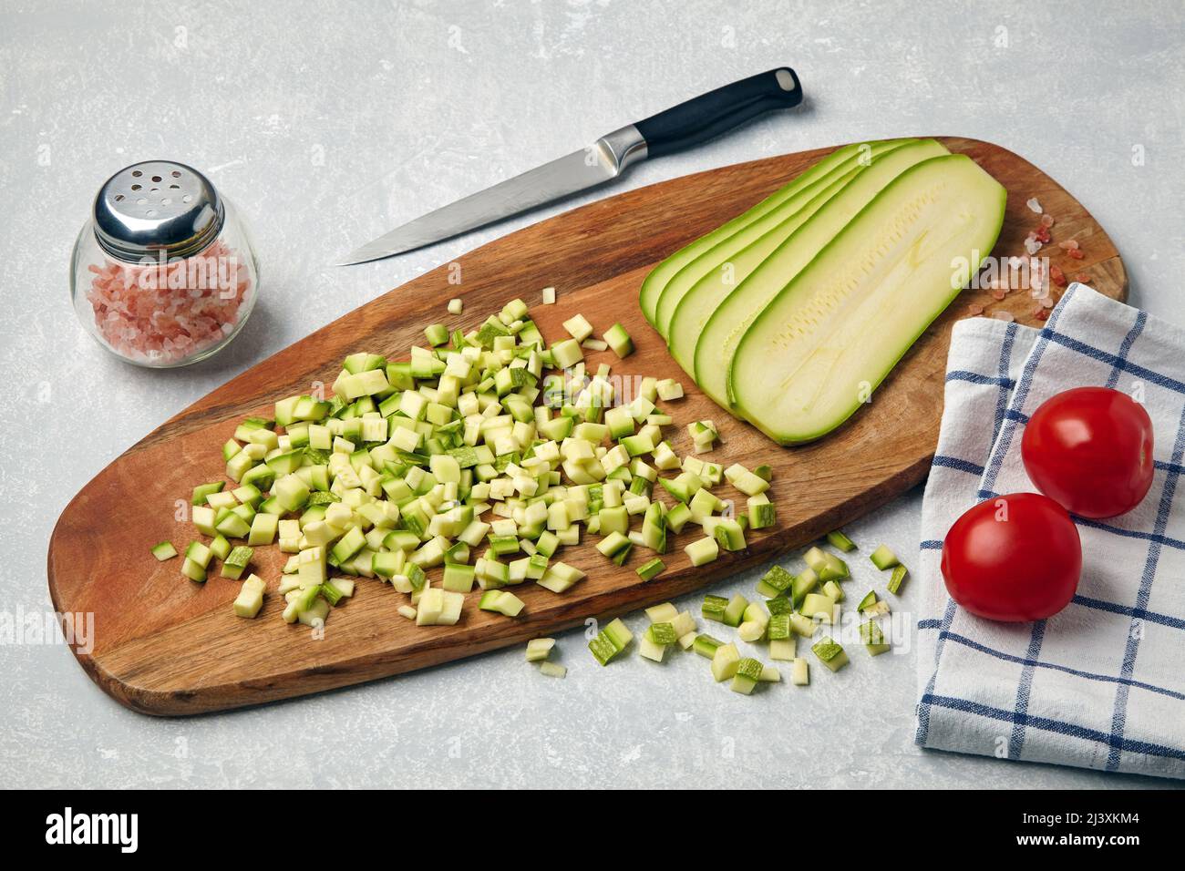 Affettato le verdure della zucchina su un tagliere di legno, pomodori, aglio, sale rosa e tovagliolo su un tavolo chiaro di cemento Foto Stock
