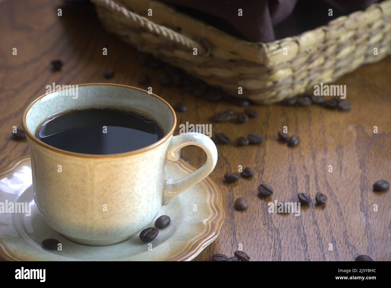 Una tazza di caffè nero su un piattino marroni con 3 chicchi di caffè. I chicchi di caffè fuoriusciti si trovano sul piano portapaziente marroni accanto a un vassoio di vimini Foto Stock