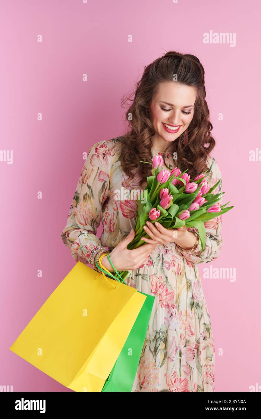 felice giovane donna in abito floreale con bouquet tulipani e borse shopping isolato su rosa. Foto Stock