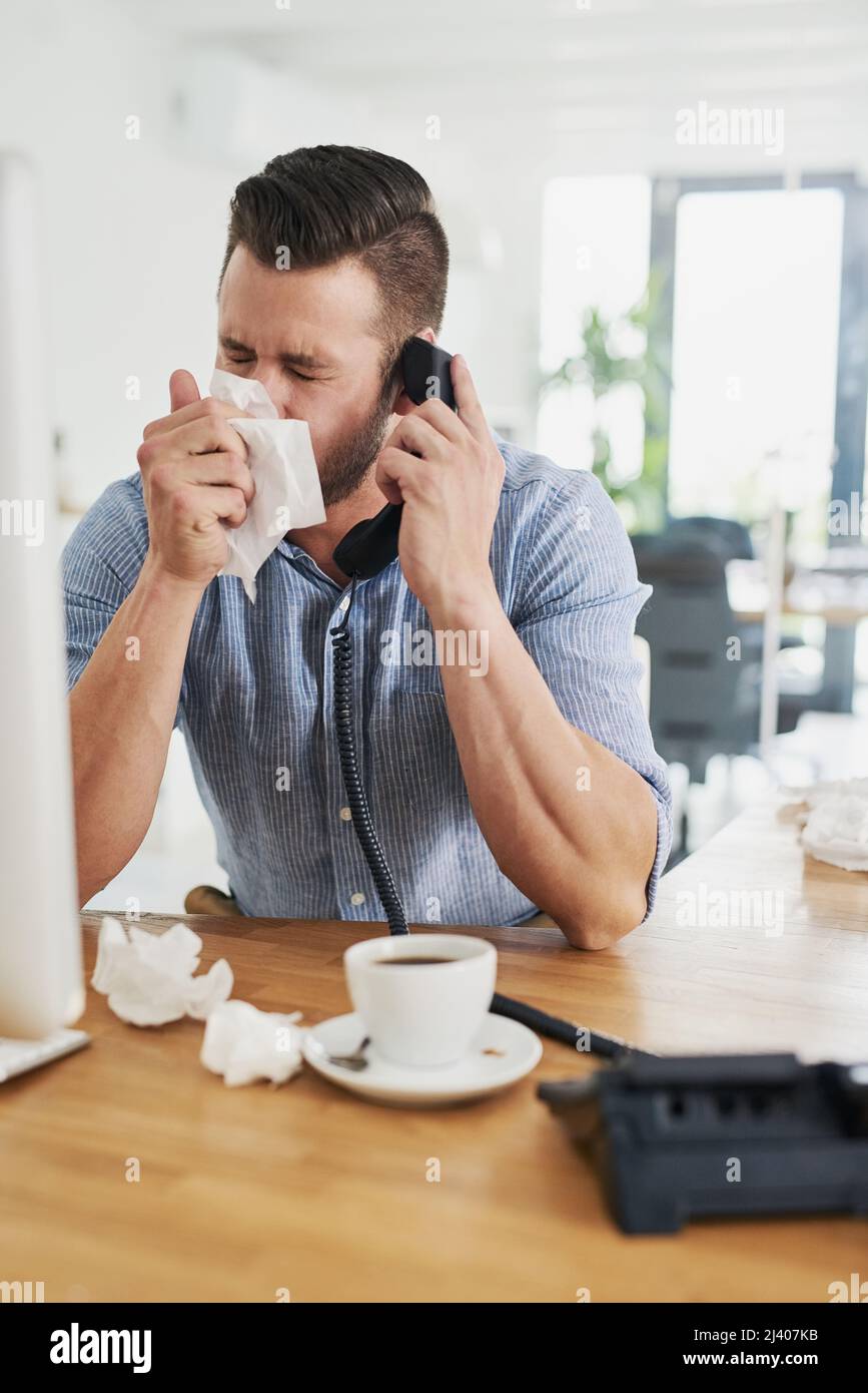 Il freddo lo ha lasciato sentire abbastanza congestionato. Scatto corto di un giovane uomo d'affari che soffia il naso mentre parla al telefono in un ufficio. Foto Stock