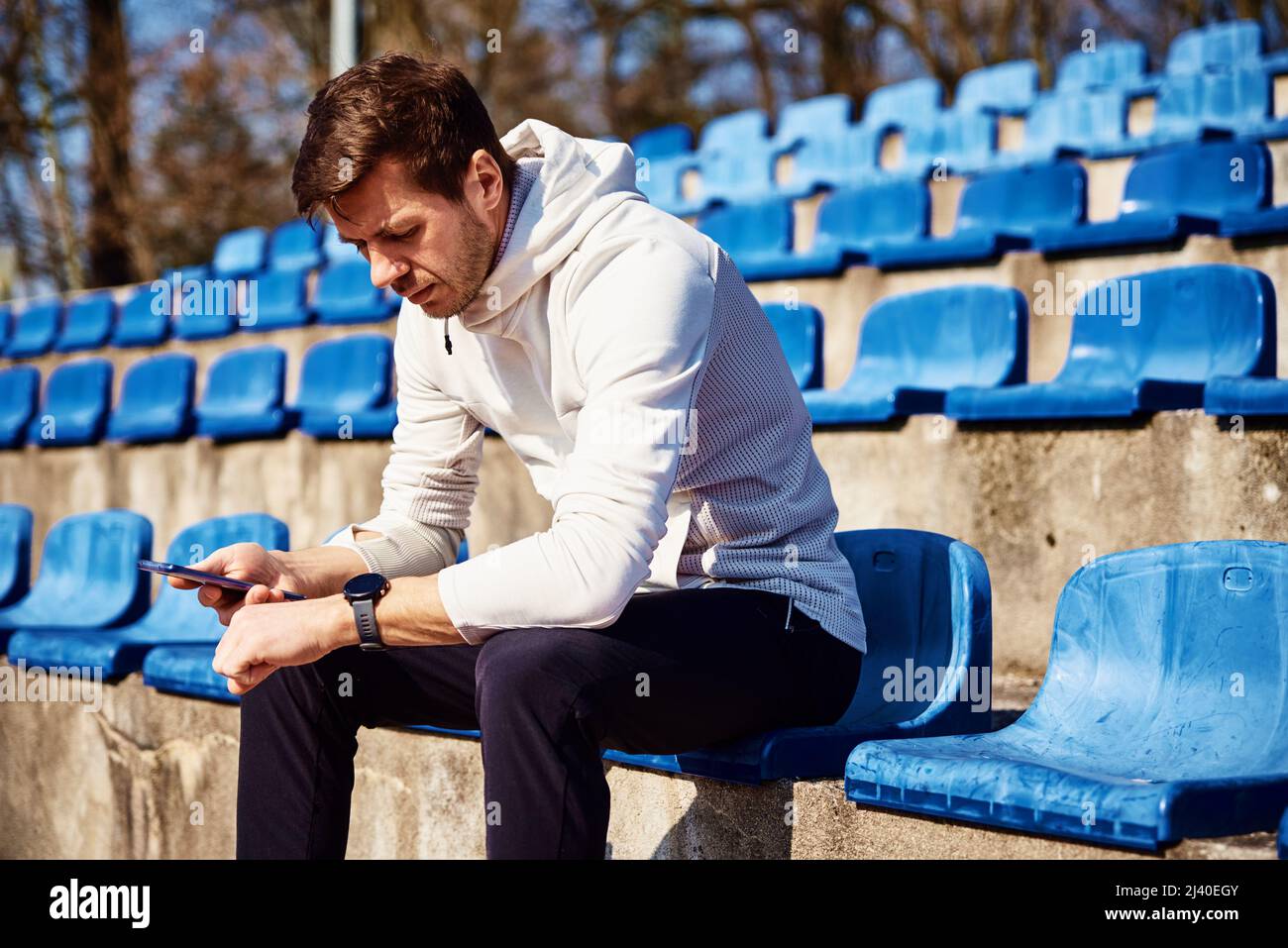 Uomo in abbigliamento sportivo seduto sul sedile dello stadio e smartphone di navigazione, uomo sportivo controllare orologio fitness dopo gli esercizi sportivi Foto Stock