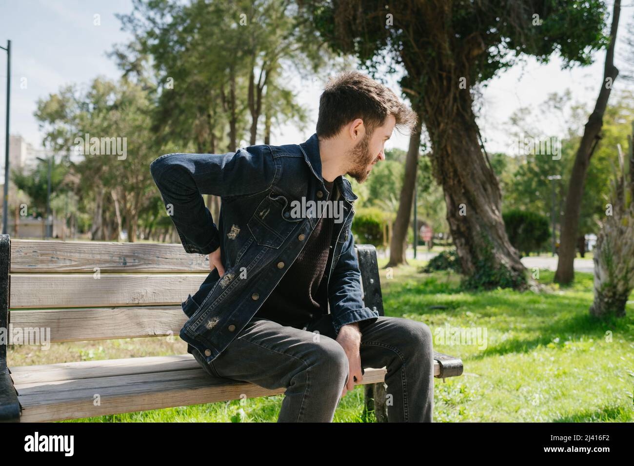 Giovane indossare jeans giacca uomo malato sensazione schiena mal seduta panca, ernia disco, problema spinale Foto Stock