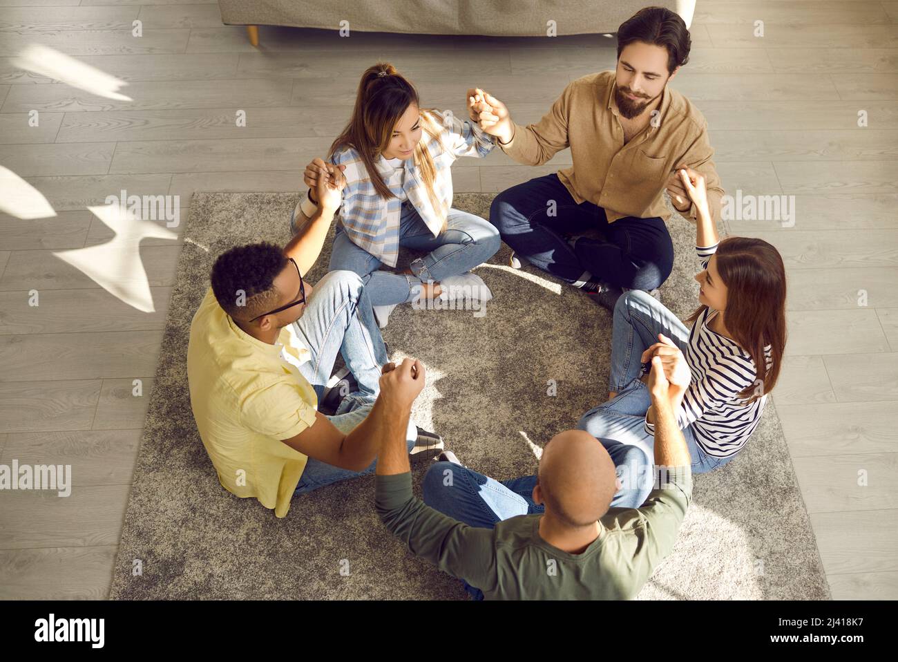 Gruppo di amici diversi seduti in cerchio sul pavimento, tenendo le mani e meditando insieme Foto Stock