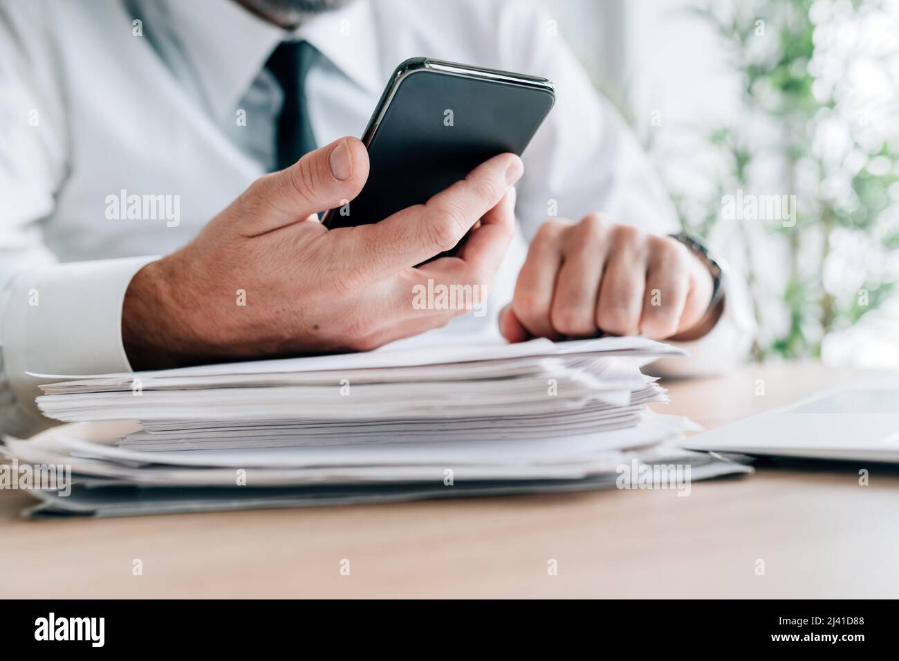 Uomo d'affari che cerca informazioni online utilizzando lo smartphone su documenti cartacei e stack di report, focus selettivo Foto Stock