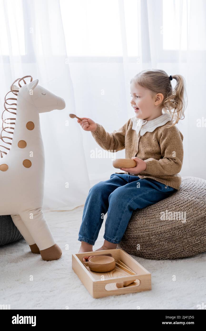 vista completa della bambina seduta sul pouf e l'alimentazione del cavallo giocattolo da una ciotola di legno Foto Stock