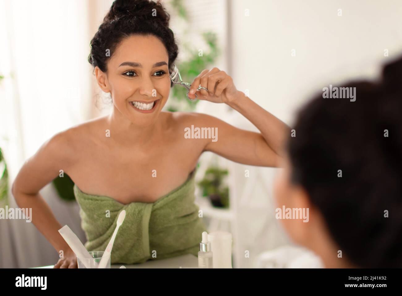 Bella giovane donna utilizzando ciglia arricciatore di fronte allo specchio a casa, spazio libero Foto Stock