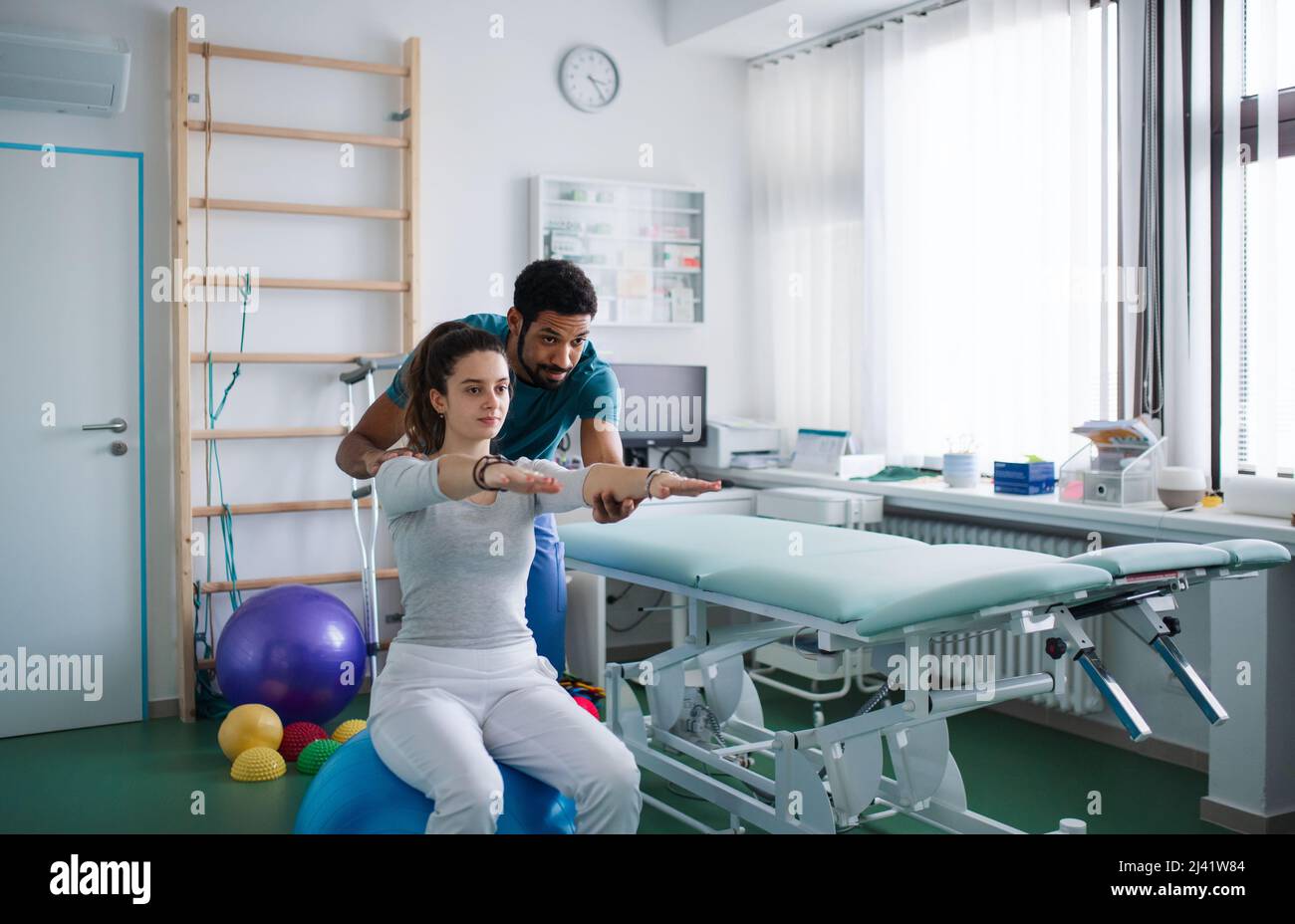 Giovane fisioterapista maschile che si esercita con giovane paziente su palla in una stanza fisica Foto Stock