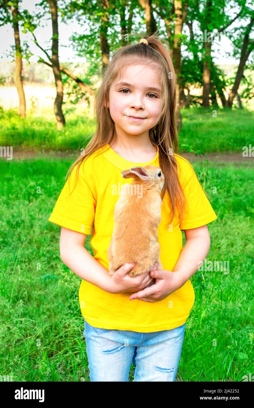 La ragazza carina piccola gioca con un coniglio di zenzero reale su uno sfondo di piante verdi. Attività estive all'aperto per bambini con animali domestici. Foto Stock