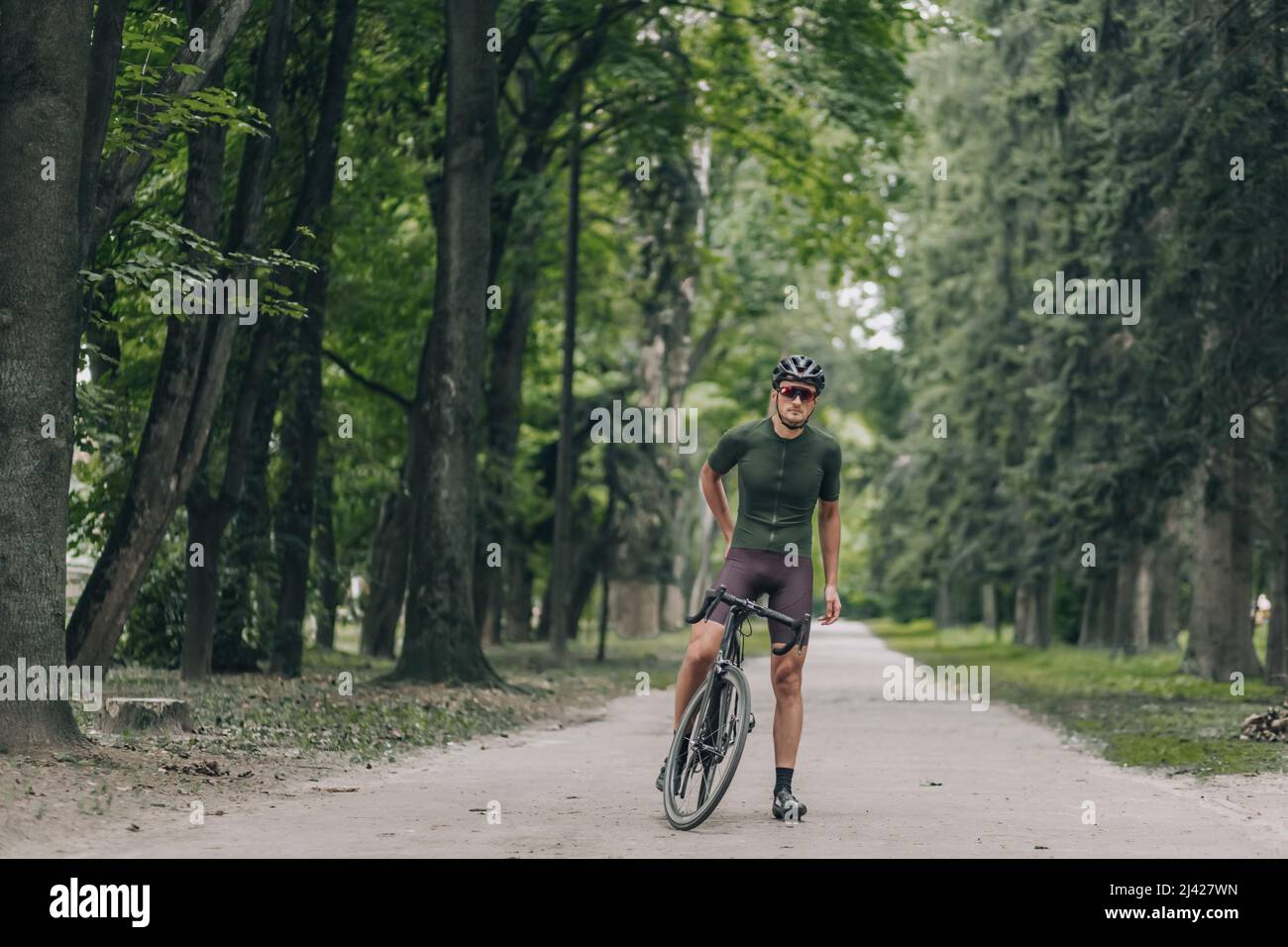 Giovane uomo attivo in piedi con la sua bicicletta nel verde parco della città. Ritratto completo di atleta rilassante durante l'allenamento all'aperto. Ricreazione e persone co Foto Stock