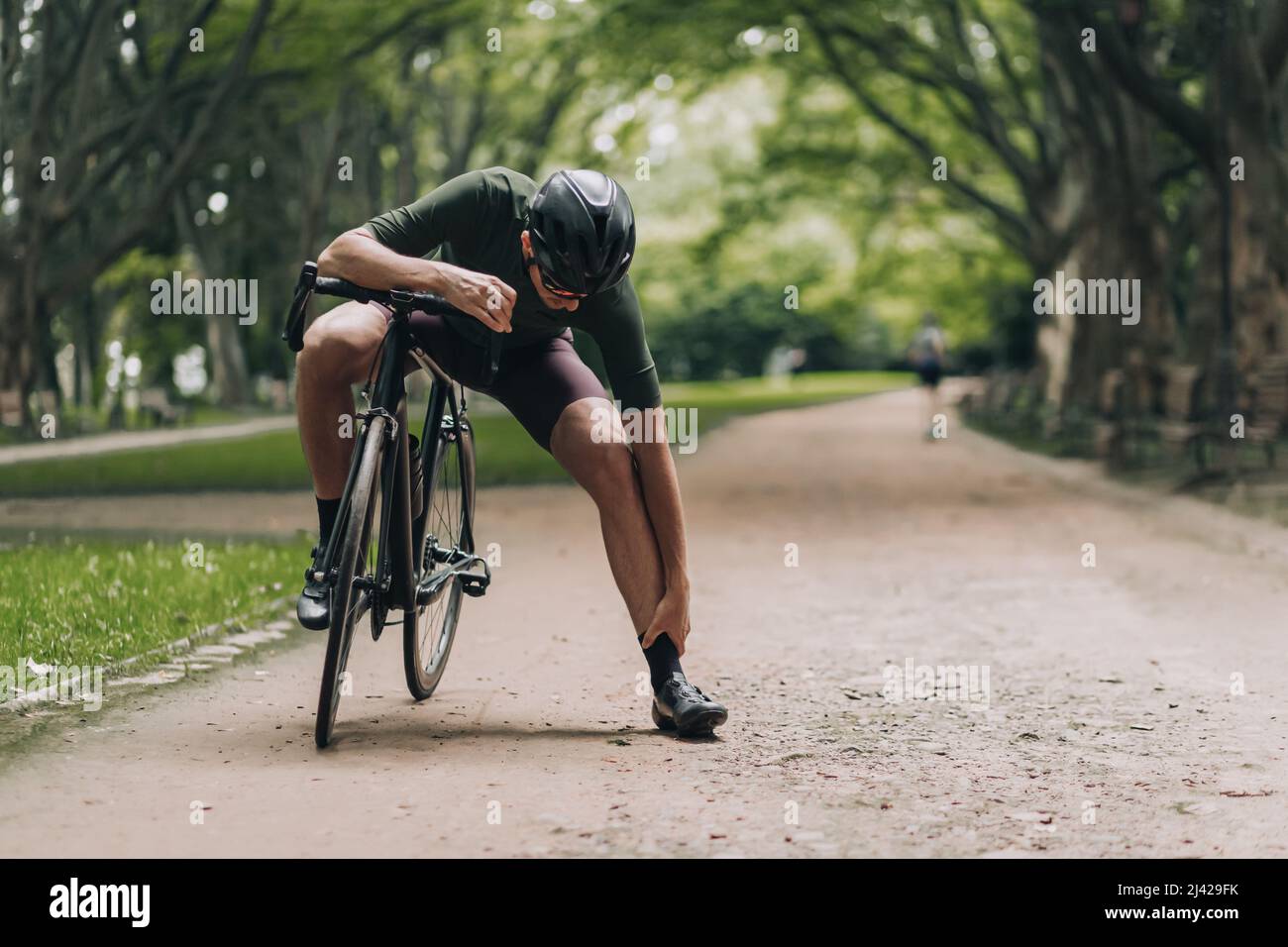 Sportivo caucasico esausto che massaggiano le gambe dopo un giro a lunga distanza in bicicletta nera. Ciclista professionale seduto al parco verde e rigenerante energia af Foto Stock
