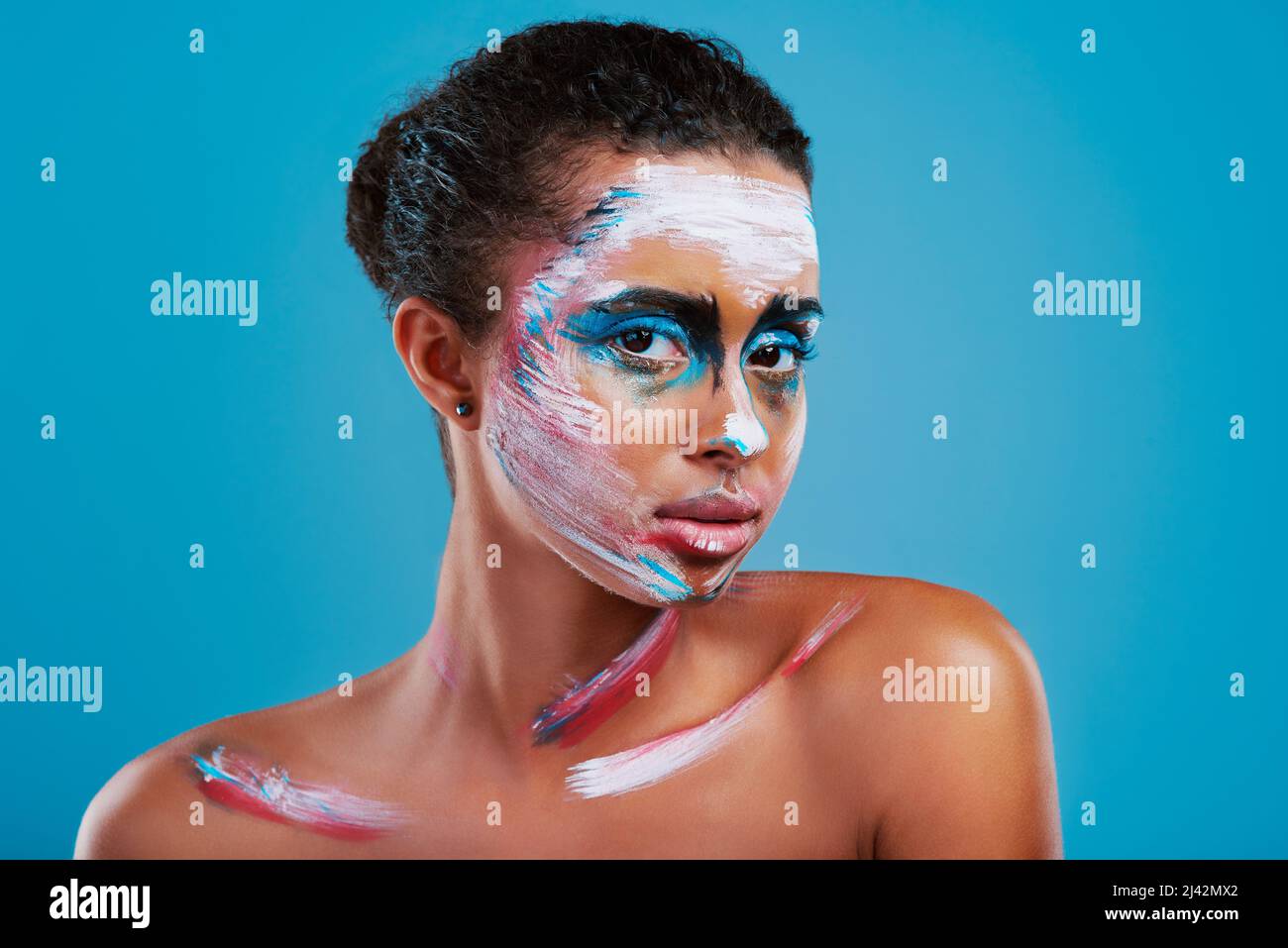 Luminoso, audace e bello. Studio ritratto di una bella giovane donna coperto di vernice facciale posando su uno sfondo blu. Foto Stock