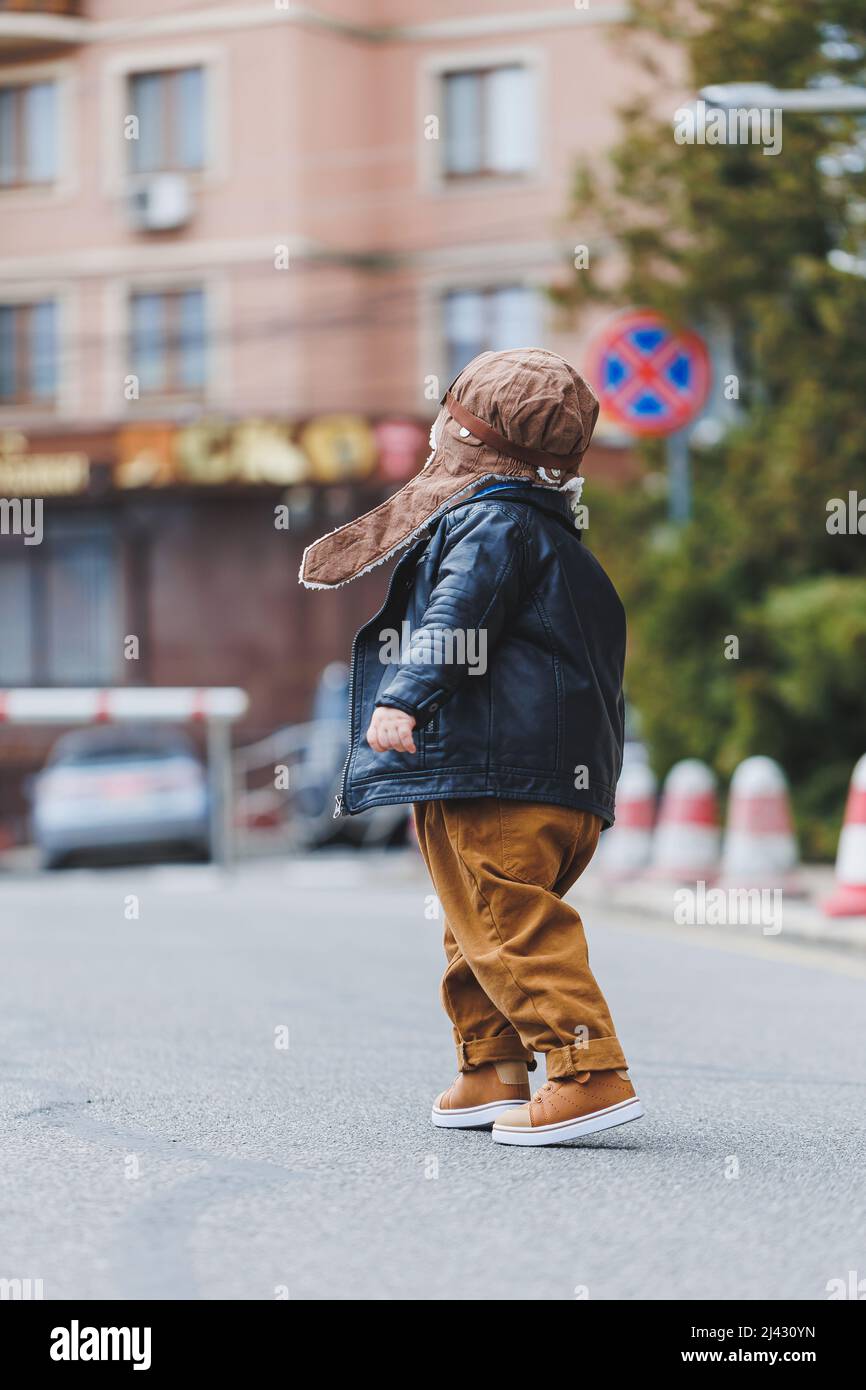 Ragazzo elegante 3 anni in una giacca di pelle e pantaloni marroni cammina per la strada. Bambino moderno. Moda per bambini. Buon bambino Foto Stock