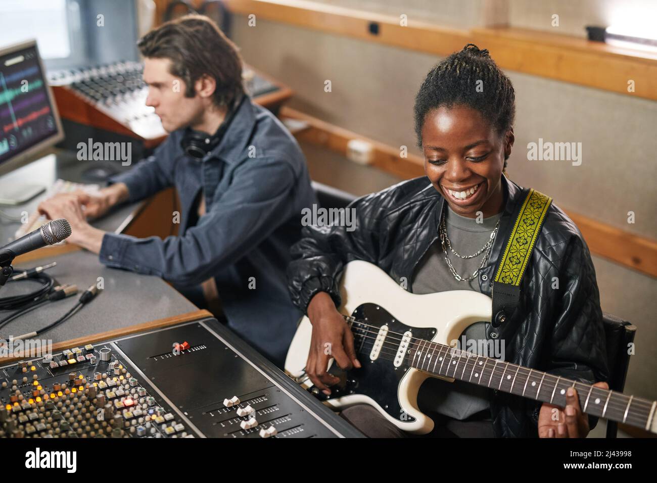 Ritratto di una donna nera sorridente che suona la chitarra mentre produce musica in uno studio di registrazione professionale Foto Stock
