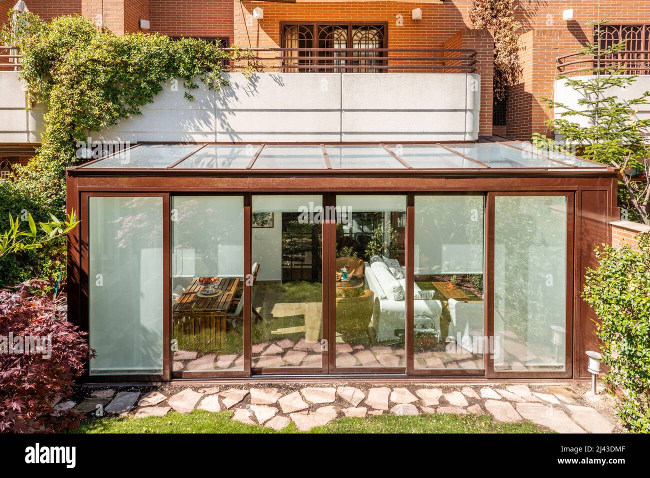 Terrazza coperta con struttura in alluminio marrone e vetro con accesso alla casa al piano terra e un giardino con un percorso di lastre di pietra e legge Foto Stock