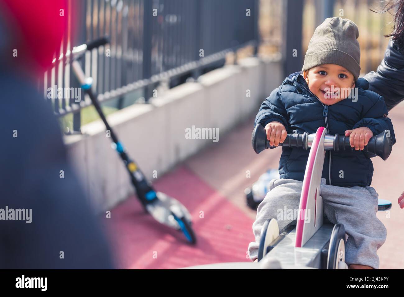 bambino felice che indossa cappotto grigio nero e pantaloni grigi seduti su una sega a vista e ridente medio pieno scatto all'aperto. Foto di alta qualità Foto Stock