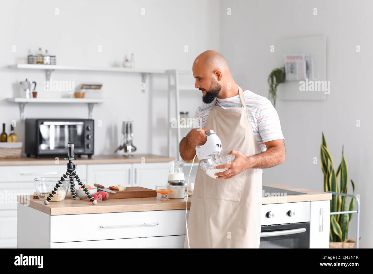 Bell'uomo che frusta l'albume d'uovo con il mixer mentre segue il video tutorial di cucina in cucina Foto Stock