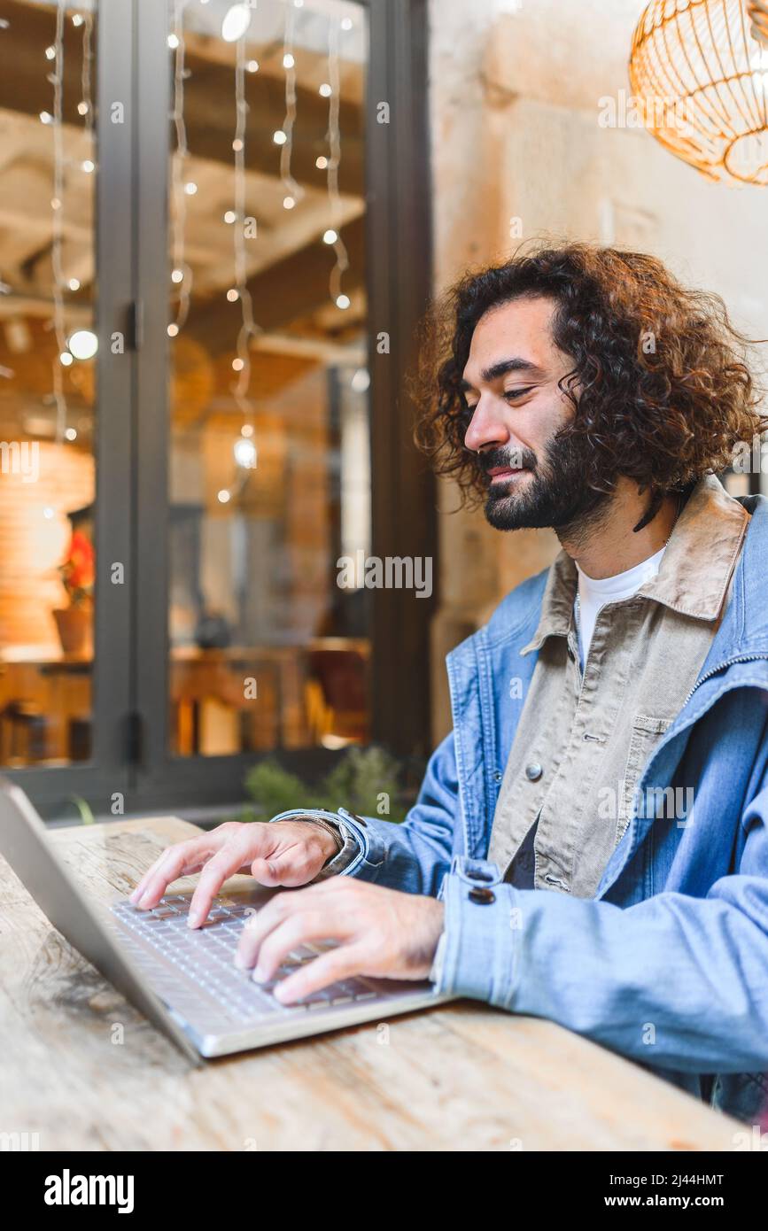 L'uomo ispanico in abiti casual con i capelli ricci digitando e leggendo i dati su netbook mentre lavora al progetto freelance in Street cafe Foto Stock