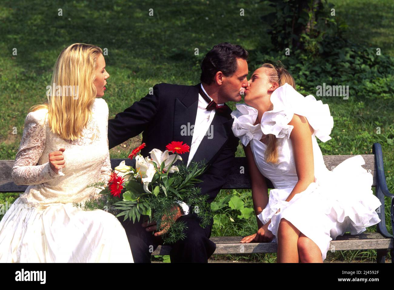 Frisch getrauter Ehemann liebt zwei Frauen, küsst seine Braut auf der Parkbank, Nebenbuhlerin schaut zu, Playboy, Vielehe, Gigola, Foto Stock
