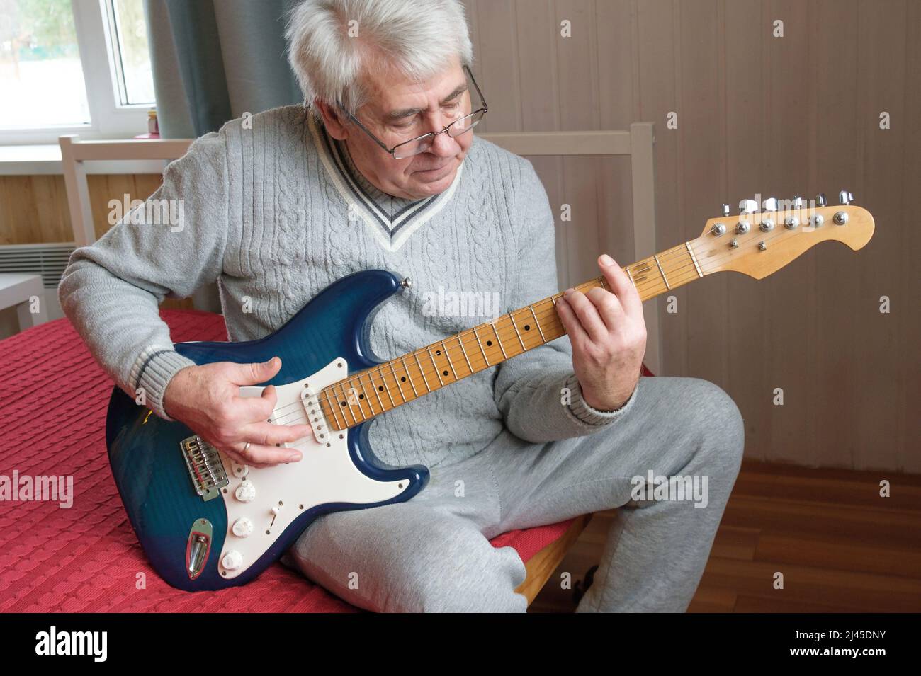 L'uomo anziano sta suonando la chitarra. Uomo anziano seduto sul divano e suona la chitarra. Ritratto di un uomo maturo dai capelli grigi in occhiali che imparano a giocare. Foto Stock