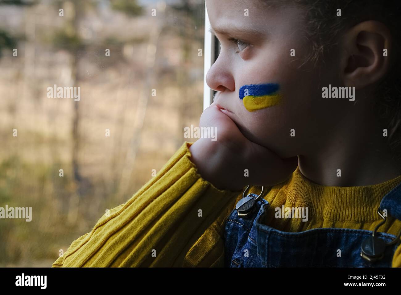 Primo piano di triste ragazzina con bandiera di Ucraina in faccia seduta da sola in windowsill a casa. Bambino infelice su sfondo naturale. Pace, nessuna guerra, lui Foto Stock