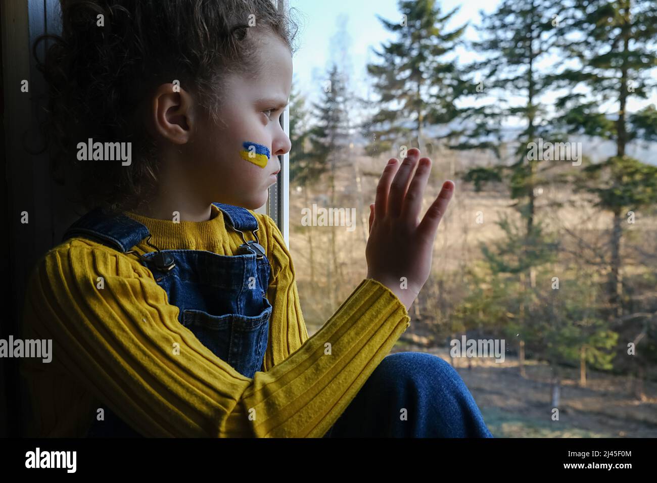 Capretto triste della piccola ragazza con la bandiera dell'ucraina sul fronte, bambino stanco infelice seduto da solo in windowsill nel paese sullo sfondo della natura. Pace, nessuna guerra, h Foto Stock