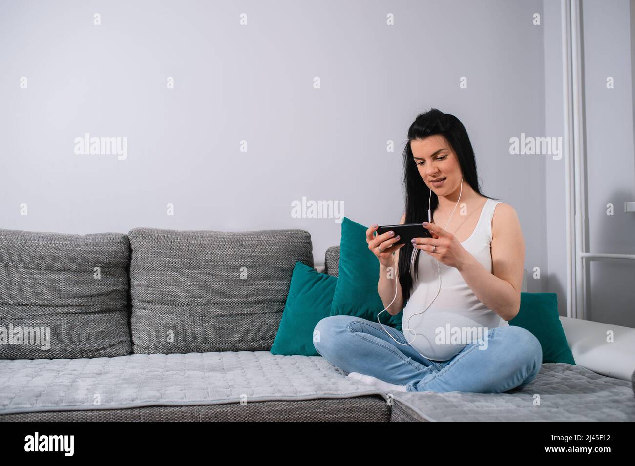 La donna incinta caucasica usa il suo smartphone e gli auricolari mentre lei si siede sul divano con le gambe incrociate nel soggiorno.Donna guarda il telefono grida. Foto Stock